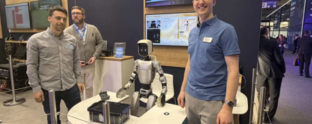 Dom Rugg-Gunn (till höger), robotikingenjör på Cambridge Consultants, vid bolagets monter på MWC 2026. Den humanoida roboten mellan teammedlemmarna är den som under mässan demonstrerade hur AI kan tolka mänskliga gester och plocka upp objekt. Foto: Nicholas Christiansen.