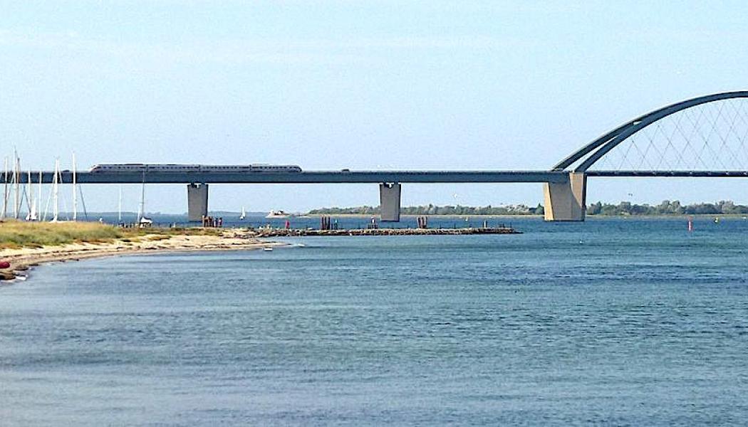 Fehmarnsundbron, vy från Großenbrode, ett tåg passerar bron. 2020 beslutade man att komplettera Fehmarnsundbron med en ny tunnel med motorväg och järnväg. Bron är K-märkt och ska behållas för lokal vägtrafik. I samband med planeringen av Fehmarn Bält-förbindelsen beslutades det att Fehmarnsundbron skulle förbli som den är, med en vägkörbana (och två filer) och ett järnvägsspår, men med elektrifiering tillagd på järnvägen. Foto: Wikipedia Common Lic. Kredit: Holger.Ellgaard - 