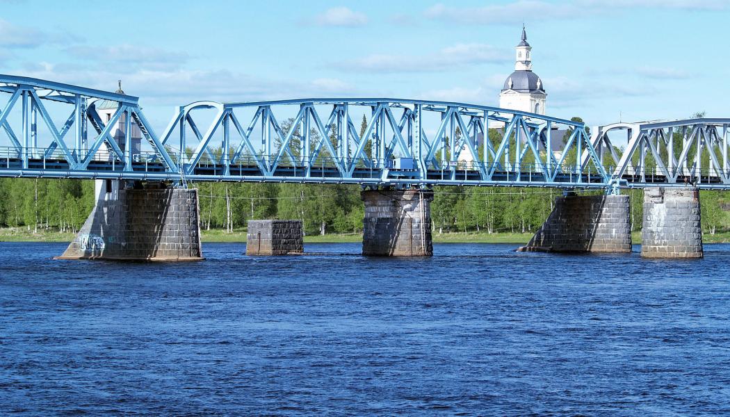 Järnvägsbron över Torne-älv. En bro mellan svenska Haparanda och den östra delen av den finska staden Torneå. Ursprungligen kunde mittspannet vändas för att låta höga skepp passera. Bakom ryttarna kyrkan i Alatornio. Foto: Wikipedia, kredit: Bobo11 